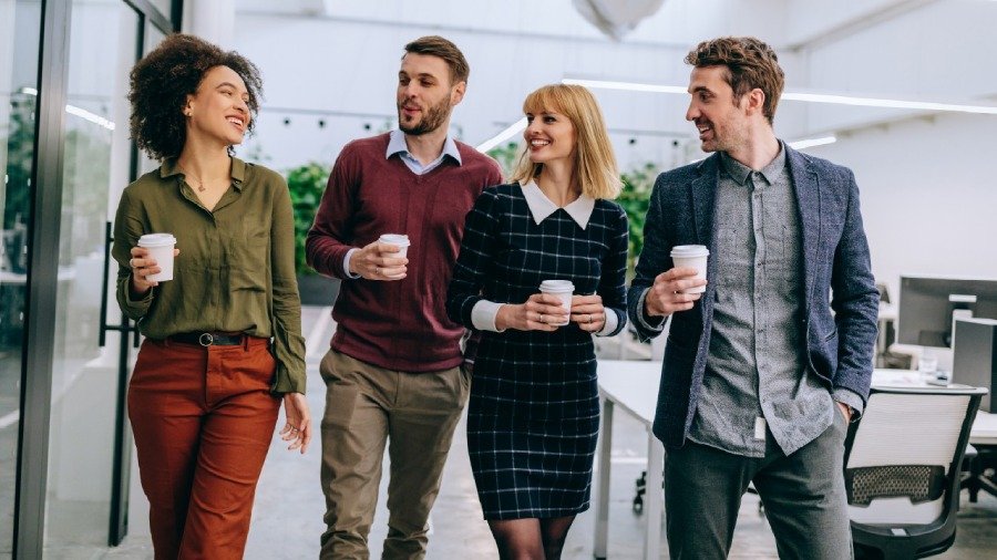 Four smiling colleagues walking together through a bright modern office holding coffee cups